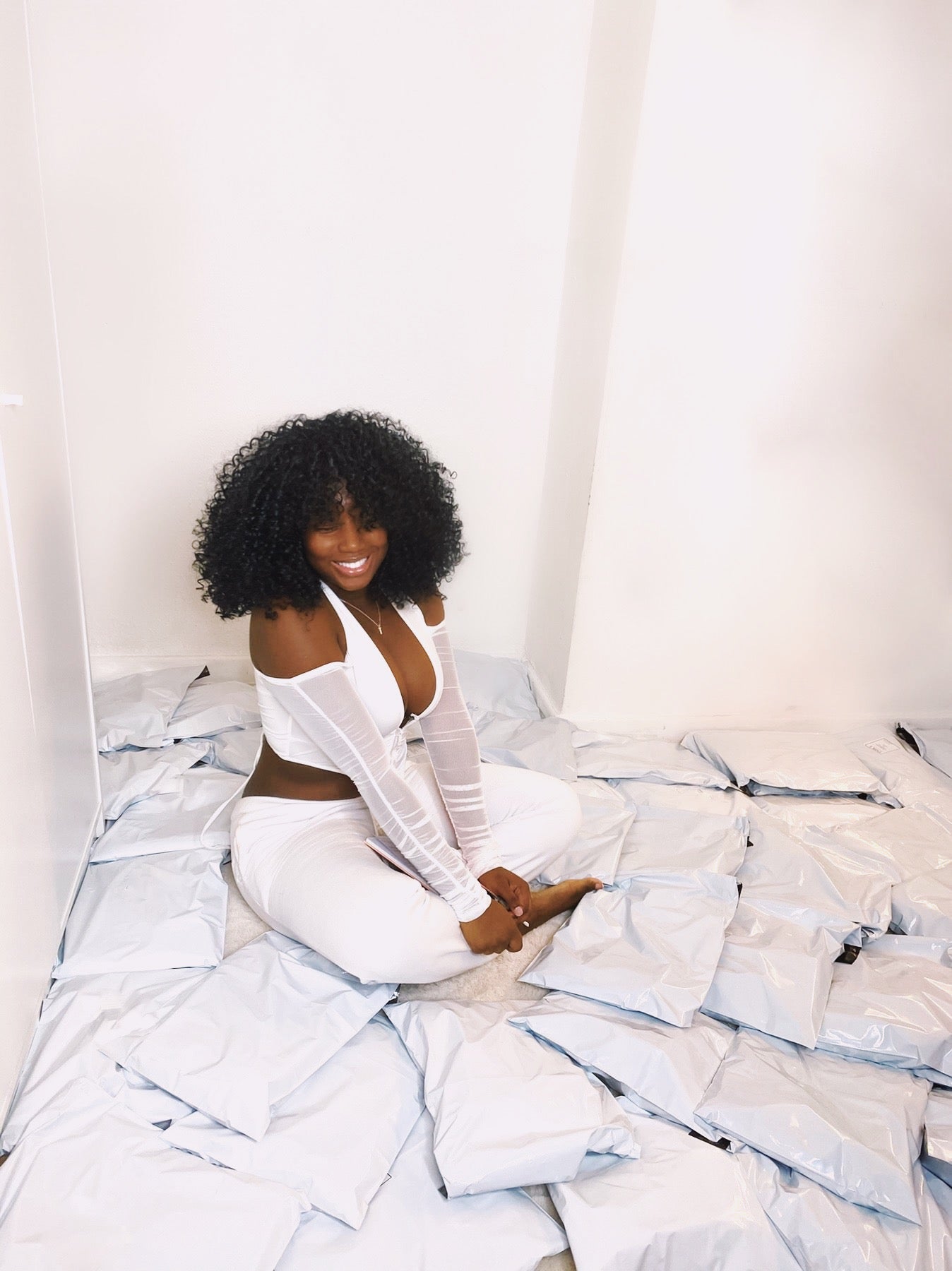 Smiling woman sitting amidst neatly packed shipping parcels, wearing a chic white outfit with curly hair, embodying confidence and style in a bright minimalist setting.