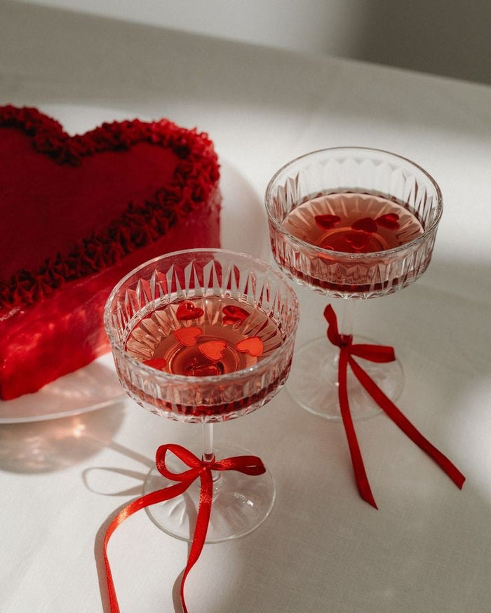 A romantic Valentine's Day setup featuring two crystal glasses filled with a pinkish-red drink, garnished with floating red heart-shaped decorations. Each glass is adorned with a red satin ribbon tied around the stem. Beside the glasses, there's a heart-s